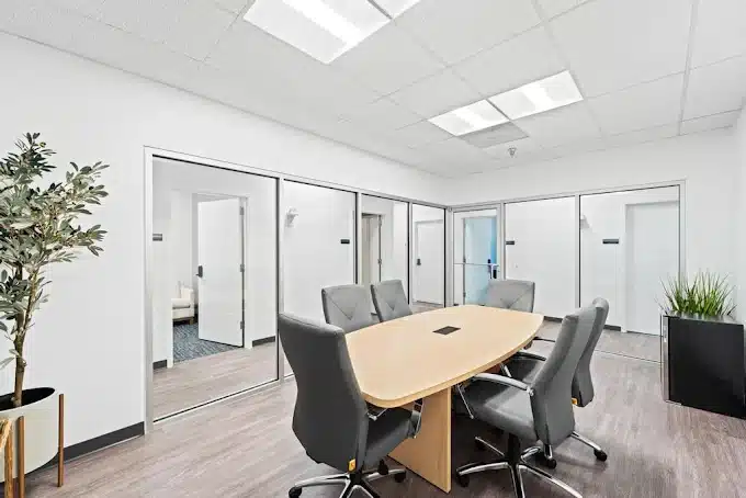 Modern conference room with wooden table, gray chairs, glass partitions, plant decor, and bright lighting.