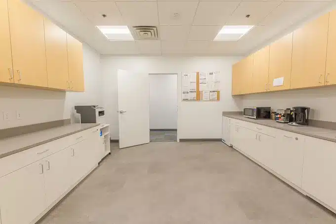 Empty office kitchen with white cabinets, beige upper cabinets, microwave, coffee makers, and open door.