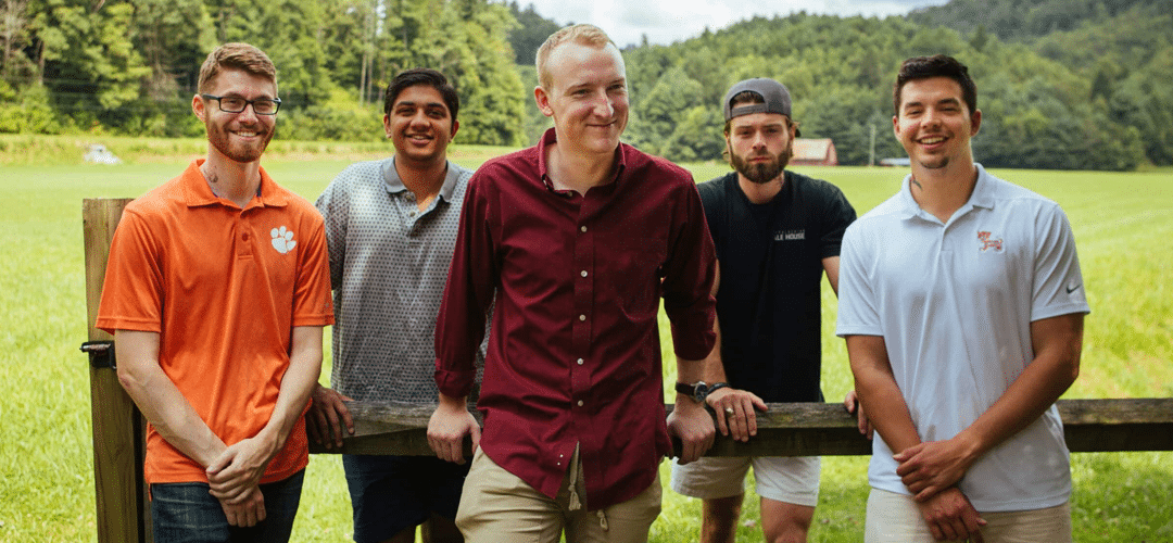Five people standing by a fence in a field, wearing casual shirts, one in red, others in orange, blue, gray, and white.