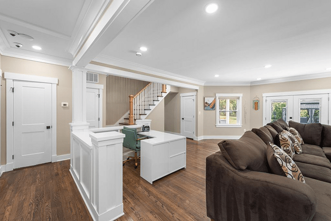 Contemporary living room with a brown sectional sofa, white desk, and wooden floors, leading to a staircase.