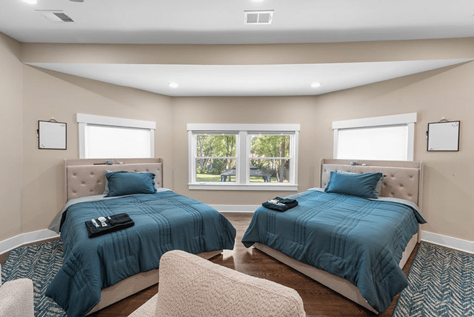 Two neatly made twin beds with blue bedding in a bright room featuring large windows and a cozy atmosphere.