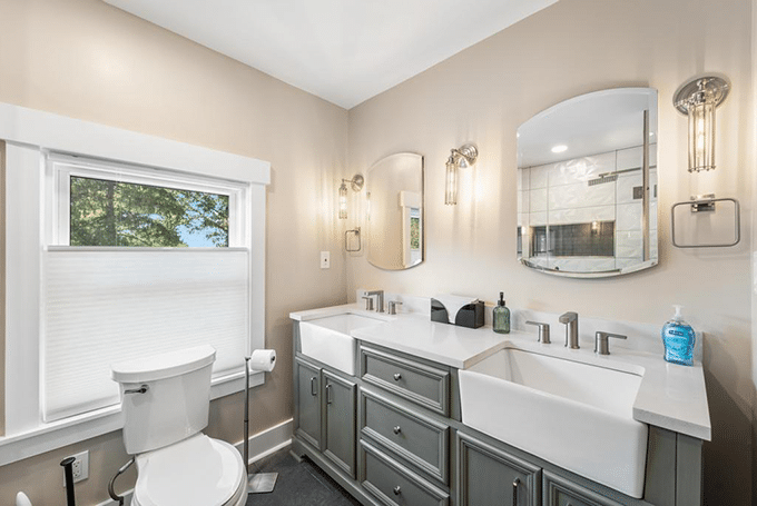 Modern bathroom with double sinks, large mirror, and natural light through a window featuring trees outside.