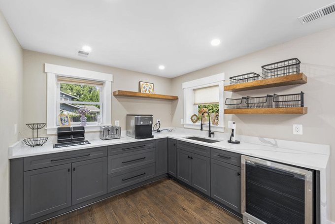 Modern kitchen with dark cabinets, open shelving, and white countertops, featuring a window view and wood flooring.