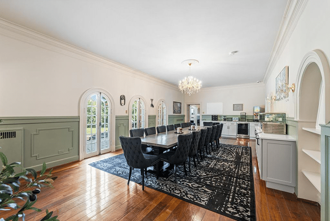 Elegant dining room with long table, plush chairs, chandelier, and arched windows overlooking a lush garden setting.