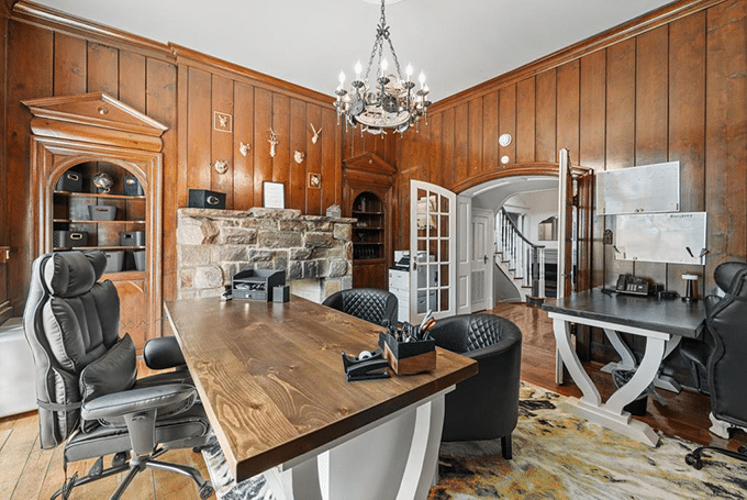 Luxurious home office featuring wooden decor, a large desk, leather chairs, a chandelier, and an elegant stone fireplace.