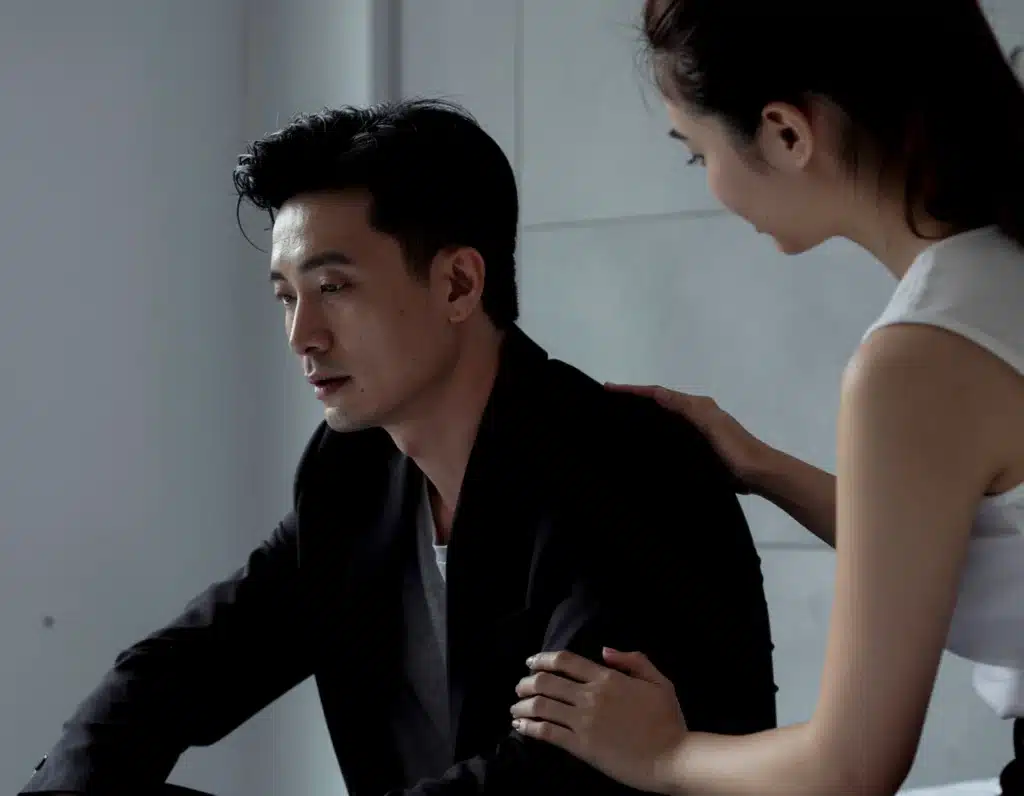 Barbiturate Addiction and Abuse: A man sitting in a dimly lit room looking sad while a woman stands at his side with a hand on his shoulder and another on his arm to provide comfort.