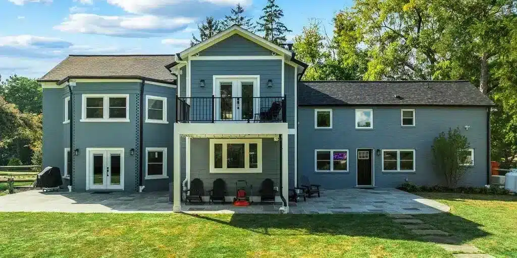 Modern two-story blue house with a spacious backyard and patio, surrounded by lush trees under a clear sky.